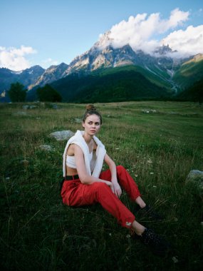 Alp tepelerinin yakınındaki çimlerin üzerinde dinlenen bir kadını gösteren açık hava sahnesi, vurgulama stili, keşif ve dağ manzarası içindeki vahşi doğa ile bir bağlantı.