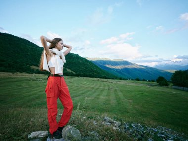 Sahne sahnesinde koyu kırmızı pantolonlu ve açık renk bluzlu bir kadın yuvarlanan tepeler ve dağlık manzaralara karşı özgürlük, seyahat tarzı ve doğa keşifleri sergiliyor..