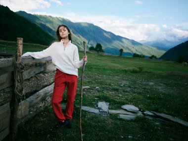 Kırsal bir arazide, bir çitin yanında duran bir kadın. Uzakta dağları olan, beyaz örgü bir kazak ve kırmızı pantolon giyen, değneği tutan, vadiye doğru bakan ve açık gökyüzüne bakan bir kadın..