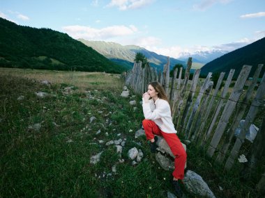 Açık hava sahnesinde yıpranmış bir çit, kayalık bir çayır ve parlak bir gökyüzünün altındaki uzak tepeler, engebeli topraklar ve refah arasındaki özgürlüğü, sakinliği ve zıtlığı yakalıyor.
