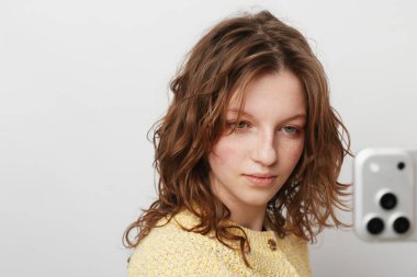 Sarı kazaklı kıvırcık saçlı kadın beyaz arka planda selfie çekerken akıllı telefon tutuyor. Yumuşak doğal ışıklandırma onun sakin ruh halini ve gündelik görünümünü arttırır. Dijital iletişim kavramı.