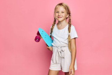 Uzun örgülü saçlı mutlu kız elinde mavi kaykayla ayakta duruyor. O, pembe bir arka plana karşı günlük kıyafetler giyer. Çocukluk kavramı, boş zaman ve aktif yaşam tarzı.