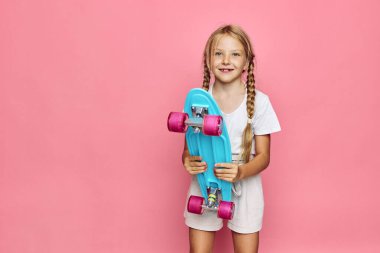 Sarı saçlı, beyaz elbiseli mutlu bir genç kız pembe arka planda duran mavi tekerlekli bir kaykay tutuyor..