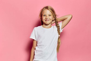 Beyaz tişörtlü mutlu kız elini başının arkasına koyup poz verirken gülümsüyor. Pembe arka plan, portre için neşeli ve parlak bir hava yaratıyor..