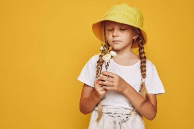 Örgülü ve sarı şapkalı çocuk dikkatlice çiçekleri tutuyor. O, gündelik hafif giysiler giyer ve sarı bir arkaplan önünde poz verir. Masumiyet, çocukluk ve doğa kavramı.