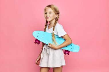 Saçları örgülü genç sarışın kız pembe bir arka planda günlük kıyafetler giyen mavi bir kaykay taşıyor. Görüntü gençliği, sporu ve modern yaşam tarzını temsil ediyor..