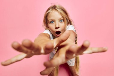 Sarışın çocuk kameraya bakarken iki eli açık ve ağzı açık bir şekilde öne doğru uzanıyor. Pembe zemin, gençlik konsepti için neşeli ve parlak bir stüdyo ortamı yaratır..