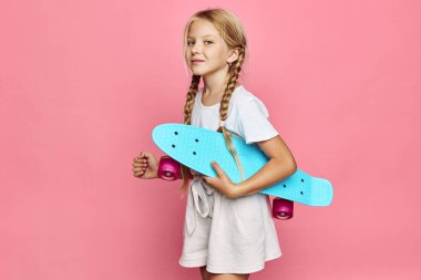 Uzun örgülü mutlu çocuk günlük kıyafetlerle mavi kaykay taşıyor. Pembe arka plan ve stüdyo ışıklandırması eğlenceli eğlence aktivitelerini ve gençlik yaşam tarzını vurguluyor.