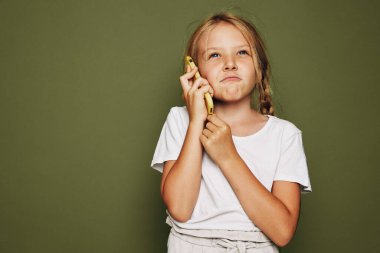 Beyaz tişörtlü meraklı kız, zeytin arka planında muz ve düşünceli bir ifadeyle telefon görüşmesi yapıyormuş gibi yapıyor. Hayal gücü ve çocukluk oyunu kavramı.