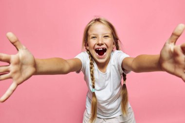 Saçları örgülü, beyaz tişörtlü mutlu bir genç kız kollarını ileri uzatıyor ve geniş bir pembe arka plan üzerinde gülümsüyor..