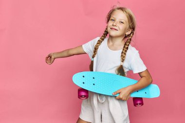 Saçları örgülü mutlu bir kız pembe duvarın önünde mavi bir kaykay tutuyor. Beyaz bir tişört ve bej şort giyiyor. Kendine güveni ve neşesiyle poz veriyor..