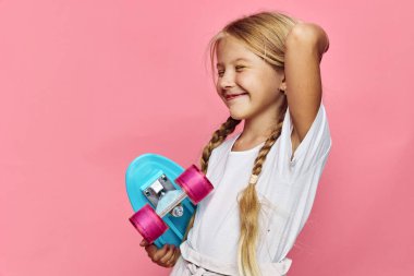 Uzun örgülü saçlı mutlu genç kız mavi bir kaykay tutuyor, günlük kıyafetler giyiyor, pembe arka planda duruyor. Yaşam tarzı, gençlik, aktivite konsepti.