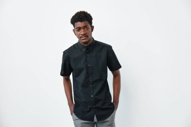 Siyah tişörtlü, siyah tenli, elleri ceplerinde beyaz arka planda poz veren Afrikalı genç bir adamın portresi..