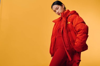 Sarı arka planda duran parlak kırmızı ceketli ve pantolonlu genç bir kadın. Kendine güvenen bir manken. Günlük kıyafetler, çağdaş tarz ve kalın kıyafetler..