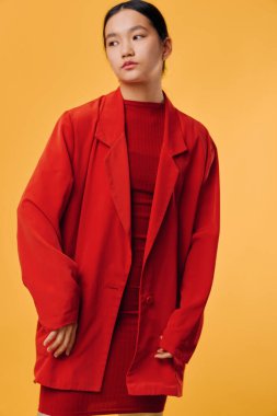 Kırmızı ceketli genç bir bayan model ve parlak turuncu arka planda dar kırmızı bir elbise. Siyah saçlı, tarafsız görünümlü şık bir kadın moda anlayışını sunuyor..