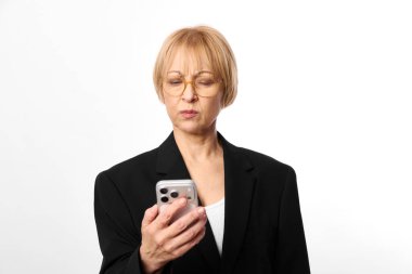 Gözlüklü ve siyah ceketli orta yaşlı kadın akıllı telefon kullanıyor. Beyaz arkaplanda yakın plan portresi. Dijital iletişim, teknoloji kullanımı, profesyonel yaşam tarzı kavramı.