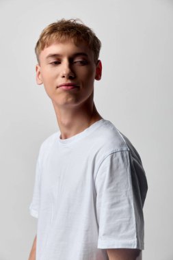 Soluk bir arka planda günlük tasarım beyaz tişört giyen rahat bir genç adam, doğal duruş ve basit ışıklandırma, rahatlık, modern moda ve çok yönlü kıyafet seçeneklerini vurguluyor.