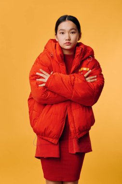 Parlak kırmızı yastıklı ceketli Asyalı kadın model ve sarı arka planda kolları çapraz duran kırmızı elbiseli. Mevsimlik kış kıyafetleri ve modern gündelik tarzıyla moda portresi..