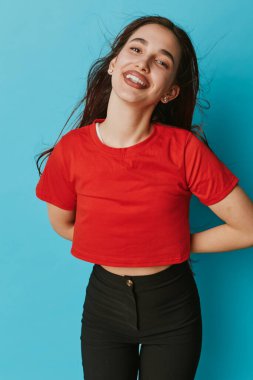 Kırmızı tişörtlü ve mavi arka planda siyah pantolonlu mutlu genç kadın poz veriyor. Çekici bir kadın, doğal bir gülüşü ve rahatlamış bir duruşu var..