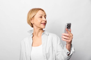 Elinde akıllı telefonu tutan son sınıf öğrencisi kadın, sakin bir ifadeyle ekrana bakıyor. Günlük beyaz giysiler ve basit arka plan teknolojiyi ve günlük yaşamı ön plana çıkarıyor..