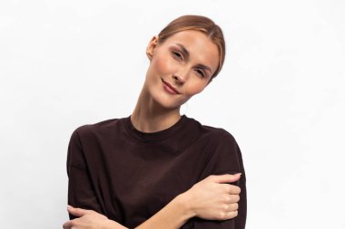 Açık tenli, kahverengi saçlı, koyu renk kazaklı, kollarını kavuşturmuş beyaz arka plana hafifçe gülümseyen genç bir kadın portresi. Güven, sakinlik, sıradan moda ve doğal güzellik kavramı..