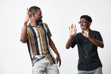 Siyah tenli, günlük gömlekli, güneş gözlüklü, dans eden ve beyaz arka planlı, pozitif ruh haliyle stüdyoda eğlenen siyahi adamlar..