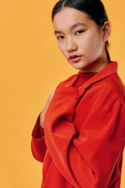 Kırmızı ceketli Asyalı kadın, parlak sarı arka planda tarafsız bir yüzle duruyor. Yakın çekim, moda fotoğrafçılığı. Stüdyo ortamında sakin bir ifade ve rahat bir poz..