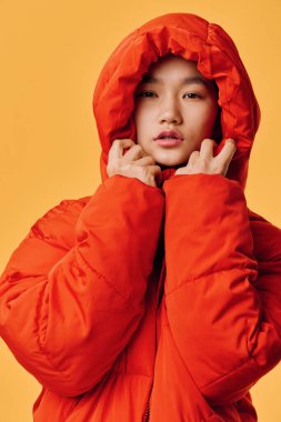 Portakal rengi kabarık ceketli kadın portresi. İki eliyle kapüşonlu. Sarı arka planlı bir stüdyo çekimi. Kış modasını ve gündelik tarzını gösteriyor. Genç bir kadında sakin bir yüz ifadesi var..