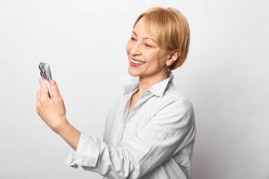 Kısa saçlı, mutlu orta yaşlı kadın beyaz arka planda akıllı telefonlu selfie çekiyor. İletişim, teknoloji, dijital yaşam tarzı ve neşeli ruh hali kavramı..