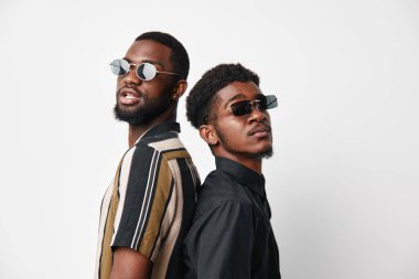 Siyah tenli, güneş gözlüklü ve günlük giysileri olan modern moda ve tavır için beyaz arka planda poz veren iki Afrikalı Amerikalı erkek portresi..