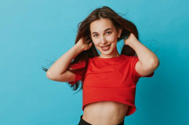 Gülümseyen, kırmızı tişörtlü, göbeği açık, elleri saçında genç bir kadın. Mavi stüdyo arka planında uzun saçlı ve rahat ifadeli bir kadın model..