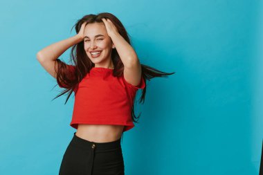 Kırmızı bluzlu, uzun saçlı ve mavi arka planda siyah pantolonlu gülümseyen genç kadın. Kadın model neşe ve güveni dışa vuruyor elleri saçlarında ve iç mekanlarda günlük tarzında..