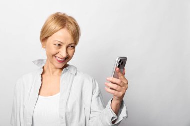 Beyaz gömlek giyen, kısa sarı saçlı mutlu orta yaşlı kadın akıllı telefonu tutuyor ve beyaz arka planda izole bir şekilde gülümseyen ekrana bakıyor. Dijital iletişim ve bağlantı kavramı.
