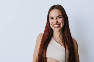 Düz uzun saçlı, beyaz kolsuz bluzlu, sade ışık duvarında poz veren gülümseyen genç bir kadın. Neşeli kadın portresi Mutluluğu ve doğal güzelliği kapalı gözlerle gösteriyor.