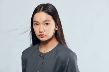 Uzun siyah saçlı, gri bluz giyen ve kameraya tarafsız bir ifadeyle bakan Asyalı genç bir kadının portresi. Temiz arkaplan ve sakin ruh haliyle stüdyo çekimi.