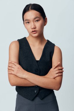 Çapraz kollu siyah yelek ve pantolon giymiş Asyalı genç bir kadın kameraya tarafsız bir ifadeyle bakıyor. Stüdyo portresi, tarz, özgüven ve basit arkaplan.