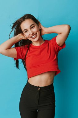 Kırmızı gömlekli, siyah pantolonlu, elleri başının arkasında poz veren bir kadın model. Rahat ve neşeli ruh halini gösteren günlük giysiler ve doğal makyajla parlak mavi bir zemin..