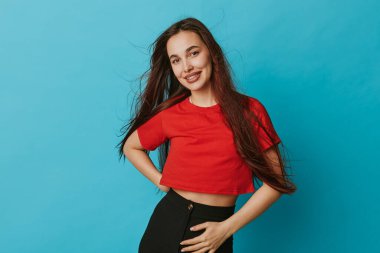 Kırmızı tişörtlü, siyah pantolonlu, eli belinde, uzun saçlı ve kameraya bakan gülümseyen genç bir kadın. Stüdyo fotoğrafı. Parlak mavi arka plan ve günlük kıyafetler..