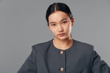 Gri arka planda düğmeleri olan gri bluz giyen sakin yüzlü genç Asyalı kadın düzgün saç stili ve minimum makyajlı kameraya bakıyor..