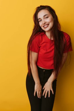 Kırmızı bluzlu ve siyah pantolonlu gülümseyen genç bir kadın elleri sarı arka planda dikiliyor. Gündelik tarz, uzun saç ve parlak stüdyo ortamında neşeli bir ifade..