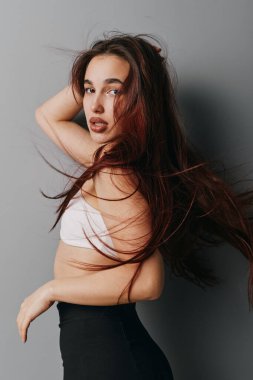 Çekici bir kadın portresi, dalgalı saçları, beyaz ekili bluzu ve siyah pantolonu var, gri duvara karşı yan poz veriyor. Model fotoğrafçılık için stil, zarafet ve modern moda..