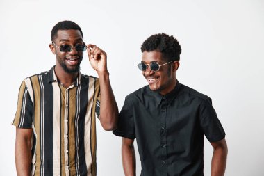 Siyah tenli, güneş gözlüklü Afro-Amerikalı erkekler beyaz arka plan stüdyosunda izole edilmiş çizgili ve siyah gömleklerle poz veriyor ve gülüyorlar..