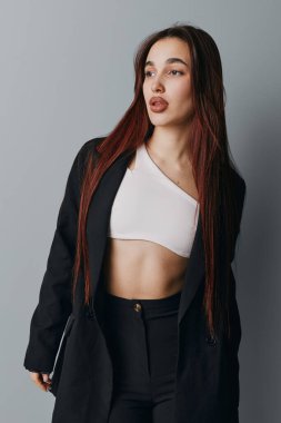 Siyah ceketli, beyaz bluzlu, modaya uygun kadın model. Tarafsız gri arka planıyla stüdyoda duruyor. Modern tarz ve kendine güveni var..
