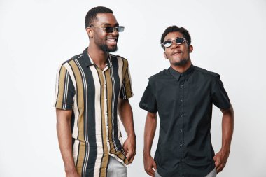 Siyah tenli, yazlık gömlekli ve güneş gözlüklü genç siyah adamlar beyaz arka planda birlikte poz veriyorlar. Afrika etnik kökeni, dostluk, moda ve rahatlamış vücut dili ile mutluluk.