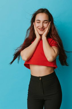 Kırmızı gömlekli ve siyah pantolonlu gülümseyen kız. Gözleri ve elleri kapalı. Yüzüne nazikçe dokunuyor..