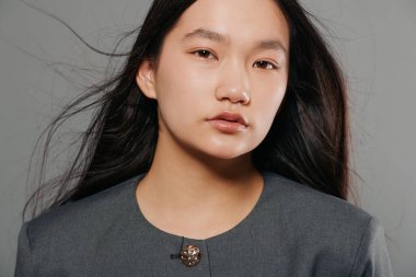 Siyah saçlı ve beyaz tenli gri elbiseli genç bir kadının portresi öne bakıyor. Tarafsız ses tonlarıyla basit bir stüdyo fotoğrafı, yumuşak ışıklandırma ve rahat bir ortam..