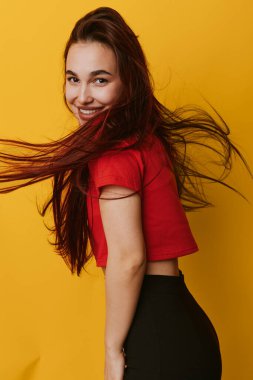 Gülümseyen, uzun saçlı, kırmızı gömlekli, siyah pantolonlu, parlak sarı arka planda genç bir bayan. Dinamik saçlı ve günlük kıyafetli neşeli bir kadının portresi..