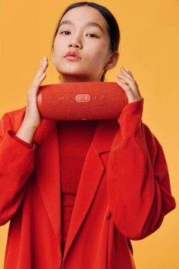 Asyalı kadın kırmızı ceket ve balıkçı yaka giyiyor. Bluetooth hoparlörünü yüze yakın tutuyor. Stüdyo çekimleri turuncu arka plan, şık bir cihaz ve son moda şehir müzik yaşam tarzı arıyor..