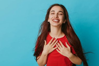 Diş teli takan, uzun saçlı, gülümseyen bir kadın. Elleri göğsünde minnettarlığını gösteriyor. Mavi stüdyo arka planında günlük giysiler içinde pozitif duygu, mutluluk ve sakin ifade.
