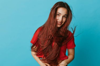 Kırmızı bluzlu, uzun saçlı, gülümseyen kız mavi arka planda duruyor. Doğal görünüş, gündelik tarz ve parlak renkler neşeli ve taze bir portre oluşturur..
